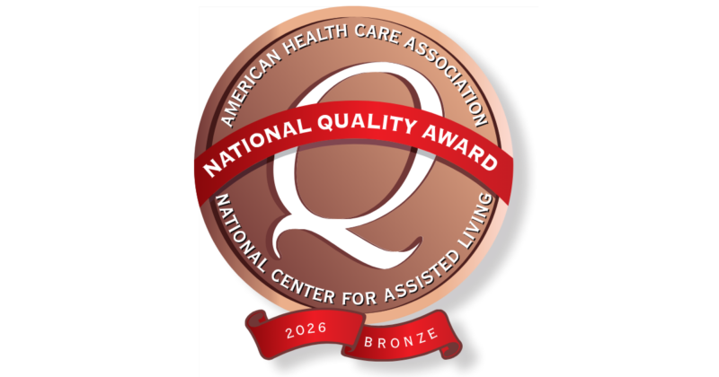 A circular bronze-colored badge with a white and red border. The outer ring reads "American Health Care Association" at the top and "National Center for Assisted Living" at the bottom. Inside, a red banner across the center says "National Quality Award." A smaller red ribbon at the bottom displays "2026" and "Bronze."